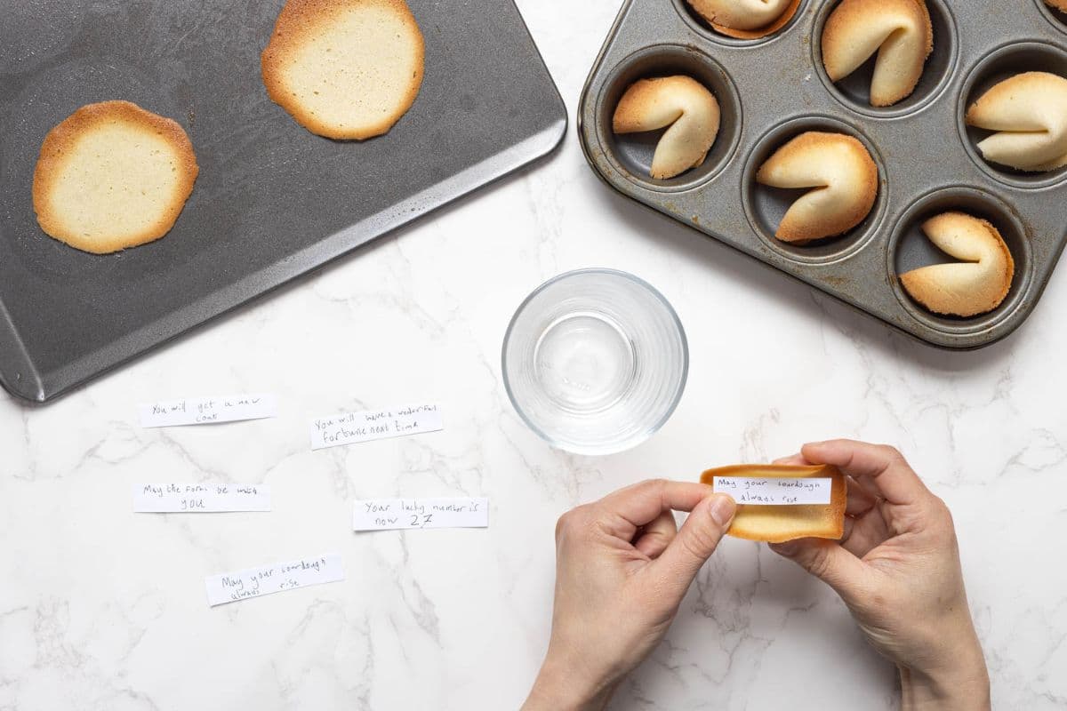 Adding hand written fortunes to the fresh made sourdough fortune cookies and shaping the cookies with a drinking glass and adding to a muffin tin.