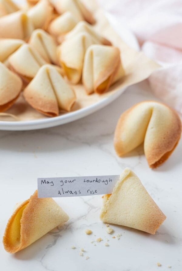 A plate of sourdough fortune cookies with one cookie broken open to show the handwritten fortune inside.