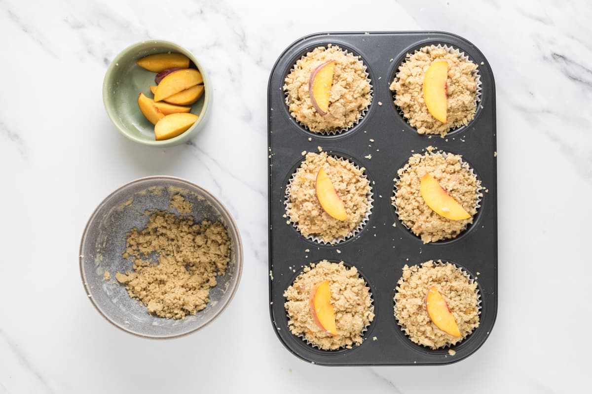Sourdough peach cobbler muffin batter in a muffin tin with homemade streusel and a peach slice on top, ready to be baked.