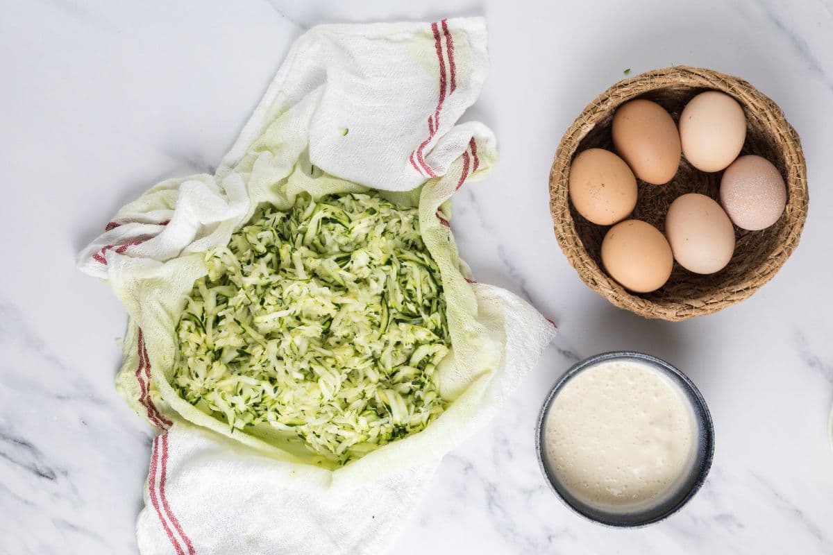 Zucchini shreds in a tea towel on the counter with sourdough discard and eggs to prepare to make a sourdough zucchini slice.