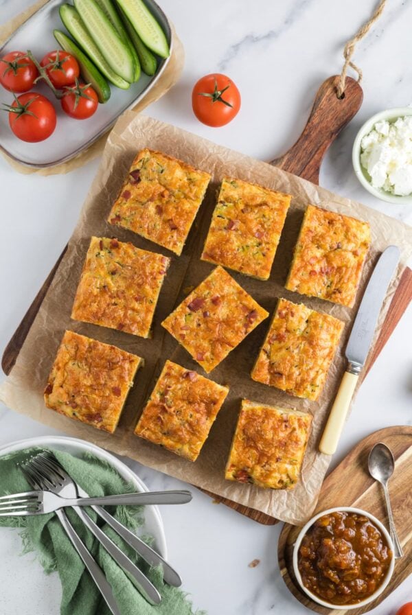 A fresh baked sourdough zucchini slice sliced on a parchment lined cutting board with condiments and vegetables.