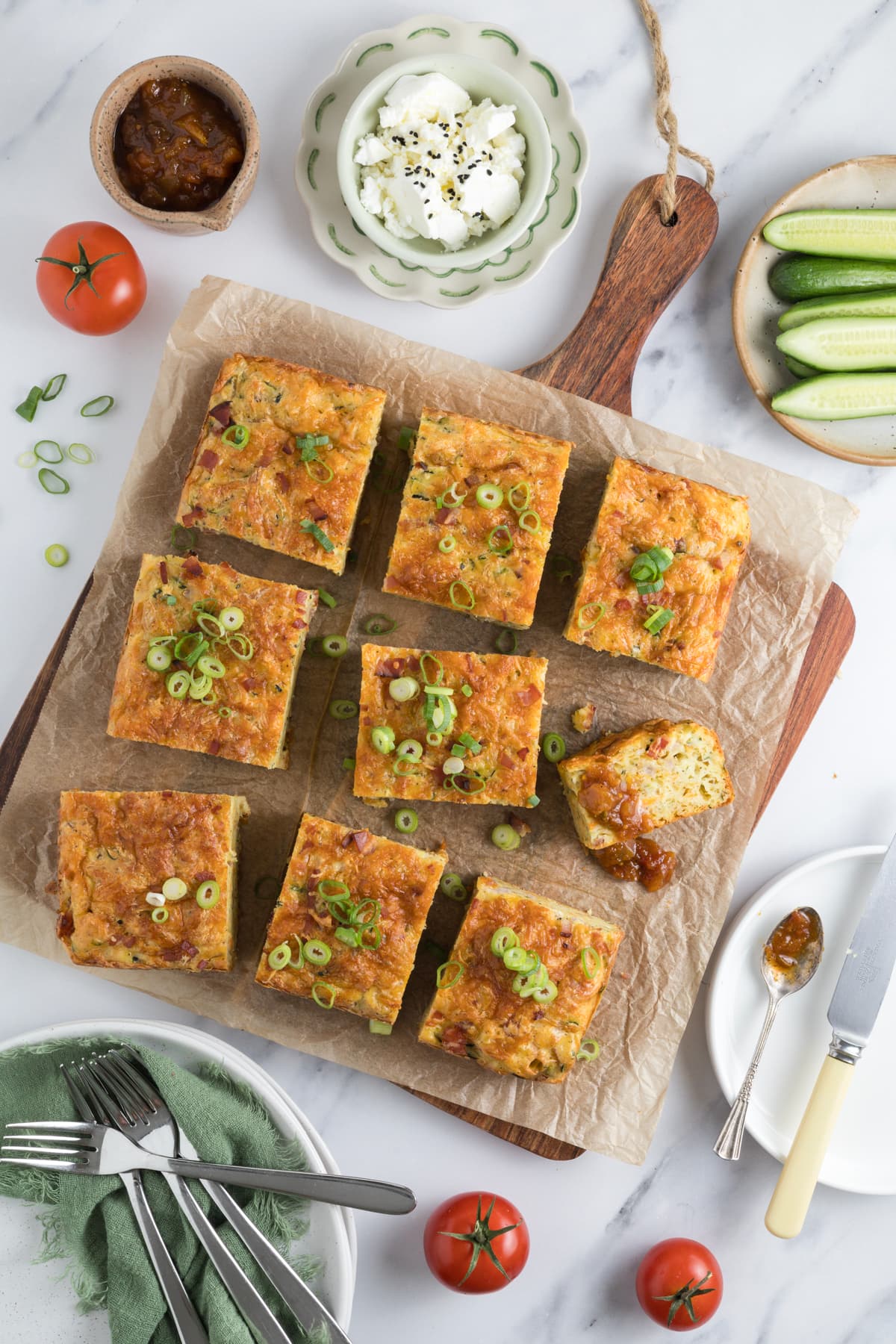 A fresh baked sourdough zucchini slice sliced on a parchment lined cutting board with spicy tomato relish on one piece and served next to fresh garden vegetables.