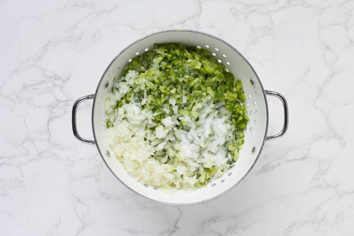 Adding vegetables to a colander with salt to drain out excess liquid to make dill pickle relish.