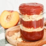 Peach cobbler jam in a glass jar with layers of buttery cookie crumbs, photographed on a wooden serving board with fresh peaches nearby.