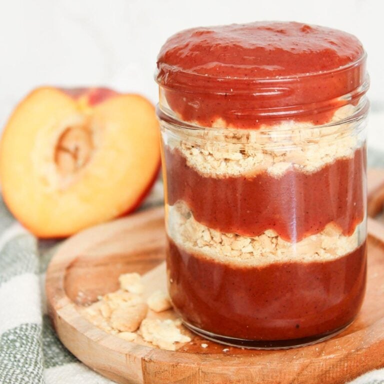 Peach cobbler jam in a glass jar with layers of buttery cookie crumbs, photographed on a wooden serving board with fresh peaches nearby.