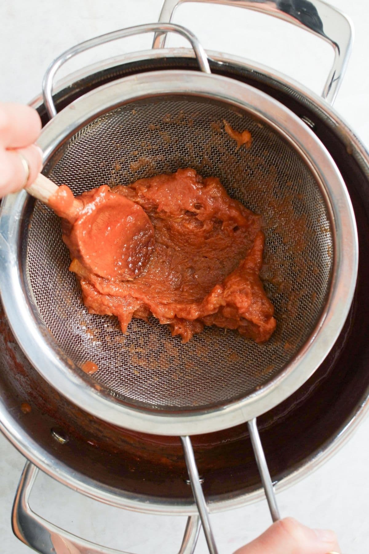 Cooked peach cobbler jam mixture being pressed through a fine mesh strainer to create a smooth jam texture.