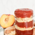 Jar of peach cobbler jam layered with crushed shortbread cookies, styled with fresh peaches and a rustic wooden board.