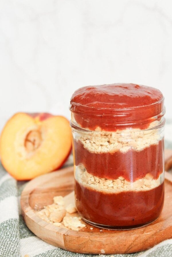 Jar of peach cobbler jam layered with crushed shortbread cookies, styled with fresh peaches and a rustic wooden board.