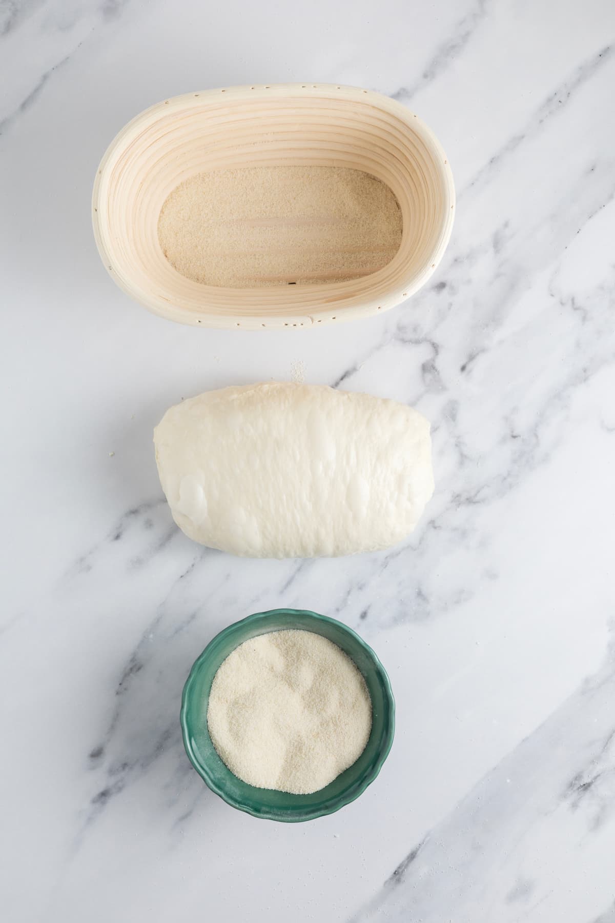 Small batch sourdough bread dough shaped on a marble countertop beside a banneton basket and semolina flour.