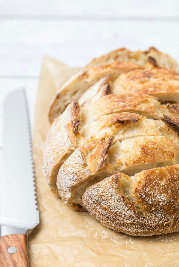 Close up of a rustic small batch sourdough bread loaf with a golden crust and deep scoring beside a bread knife.