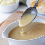 Close up of creamy sourdough gravy dripping from a spoon into a serving dish.