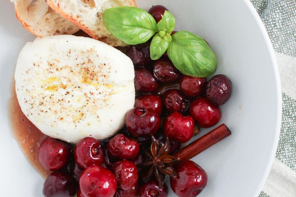 Bowl of burrata topped with sweet pickled cherries, fresh basil, sourdough bread, and spiced cherry brine.