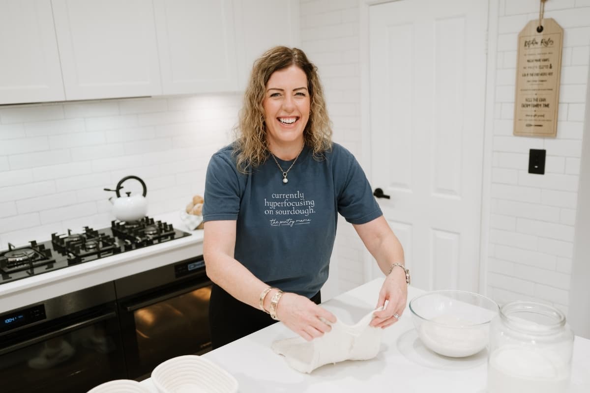 Photo of Kate Freebairn, founder of The Pantry Mama in her kitchen wearing a blue t-shirt. She is shaping a loaf of sourdough bread in her kitchen.