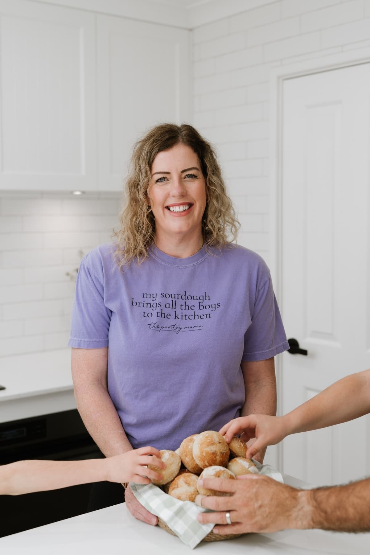 A photo of Kate Freebairn, founder of The Pantry Mama wearing a purple t-shirt and holding a basket of sourdough bread rolls. There are 3 hands reaching into the basket to take a rolls.