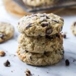 Three bakery-style protein sourdough chocolate chip cookies stacked on a marble surface with visible oats and chocolate chips throughout the cookies.