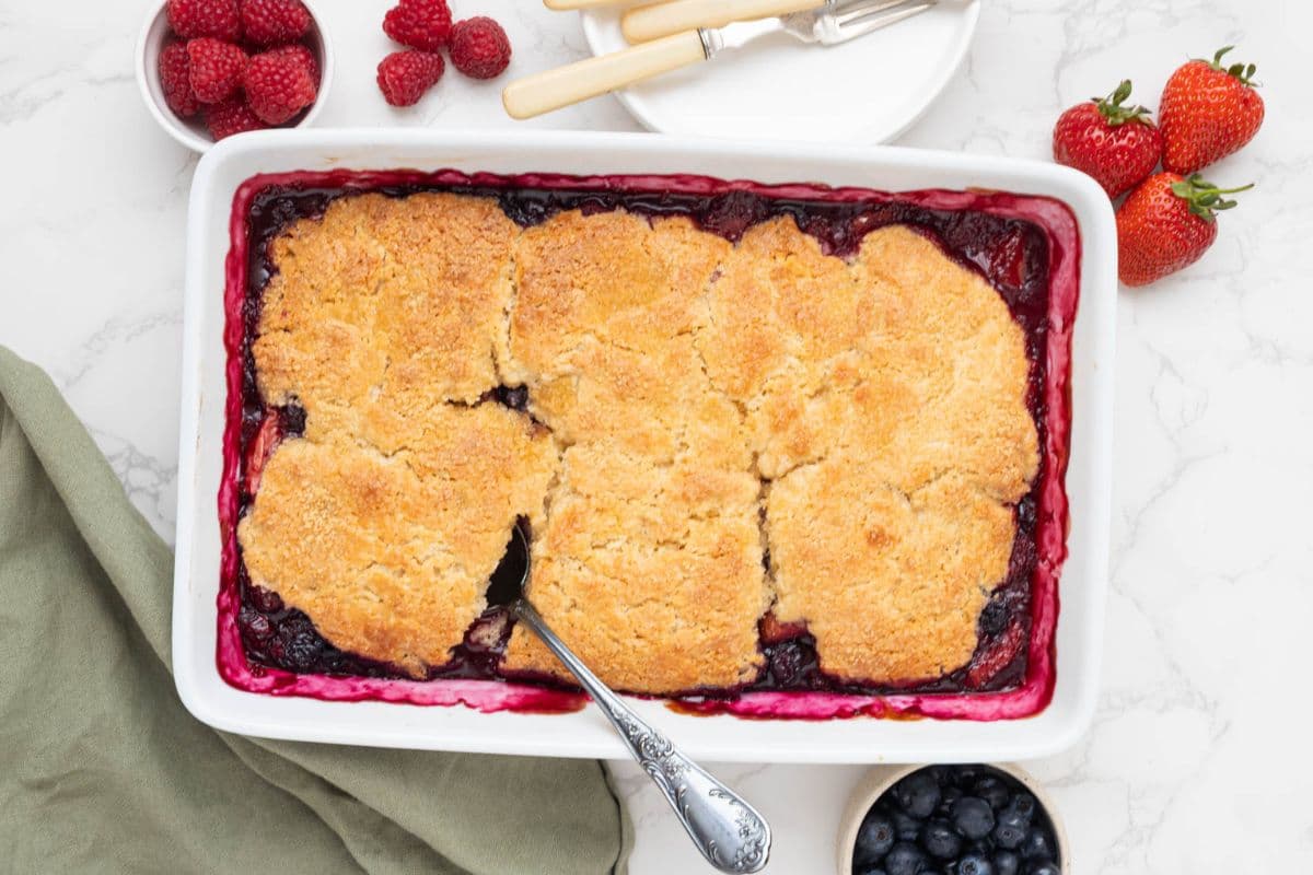 Freshly baked sourdough berry cobbler in a baking dish with a spoon inserted into the cobbler topping.