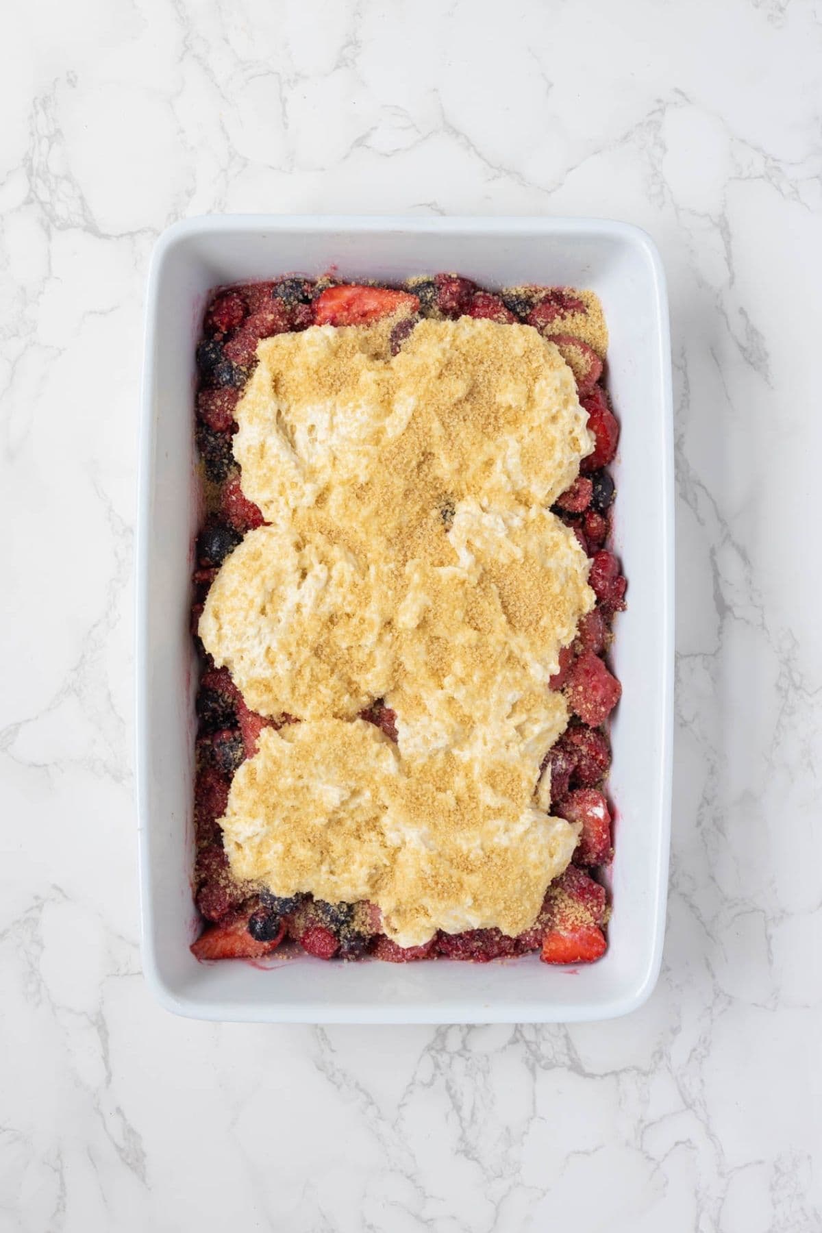 Sourdough cobbler topping spooned over mixed berry filling with sugar on top before baking.