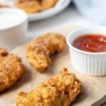 Crispy sourdough fried chicken fingers served on parchment paper with a side of ketchup for dipping.