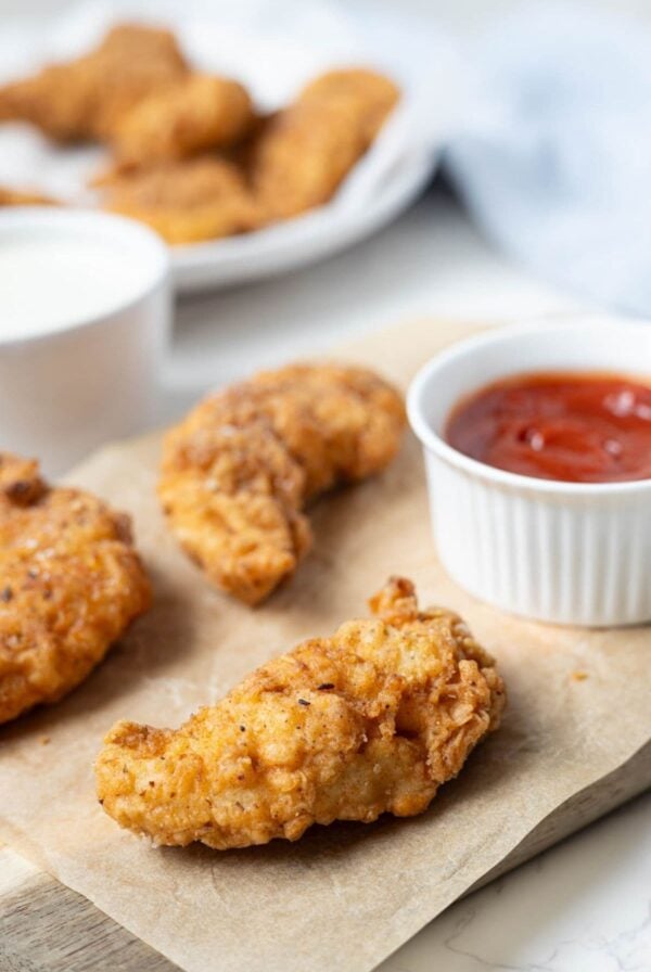 Crispy sourdough fried chicken fingers served on parchment paper with a side of ketchup for dipping.