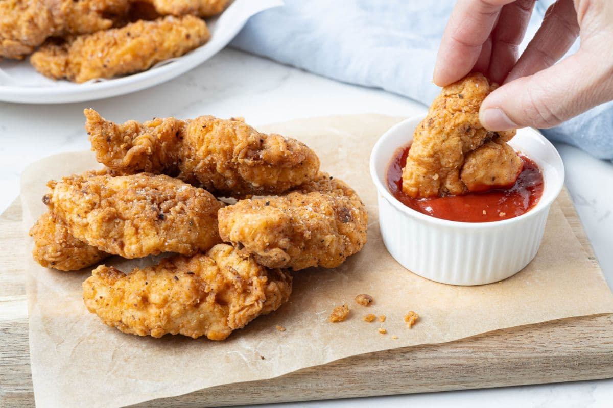Hand dipping a crispy sourdough fried chicken tender into ketchup beside a pile of golden fried chicken fingers on a wooden board.