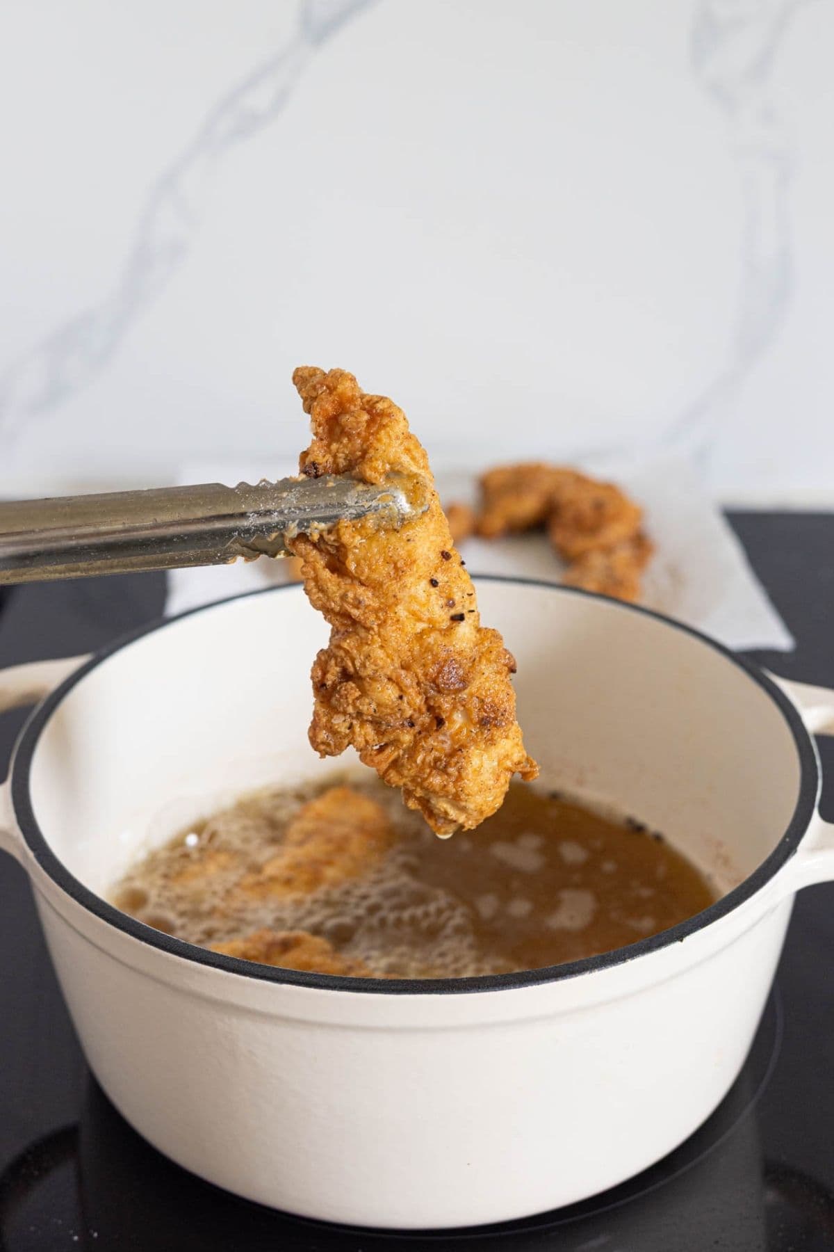 Tongs lifting a freshly cooked piece of sourdough fried chicken from hot oil in a Dutch oven.