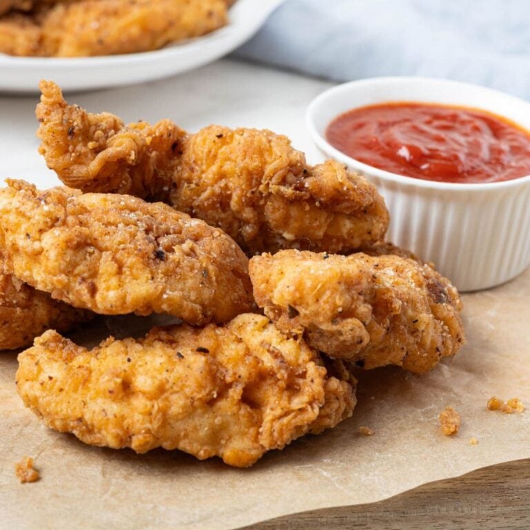 Golden brown sourdough fried chicken fingers piled on a wooden board beside a ramekin of ketchup.