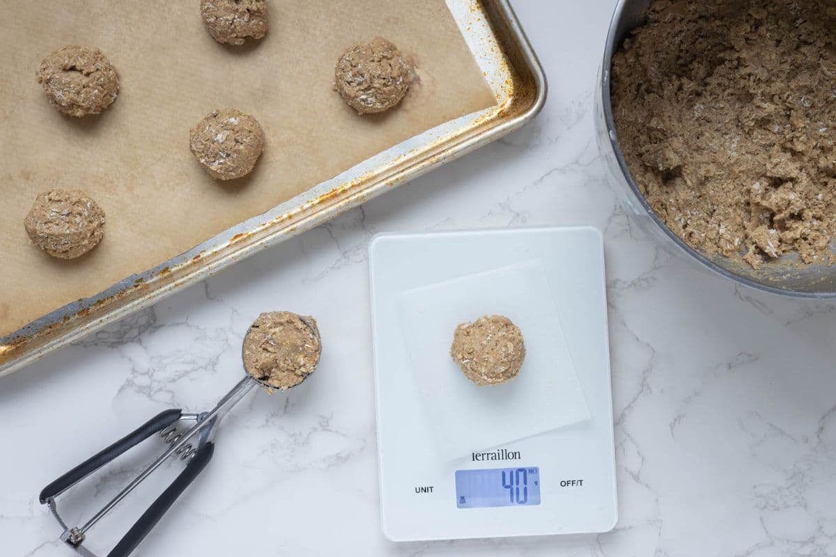 Cookie scoop portioning oatmeal cookie dough onto a baking tray with one dough ball on a kitchen scale.