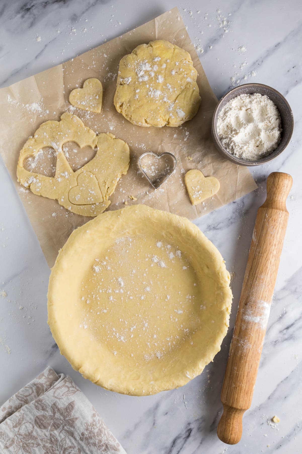 Sourdough pie crust in a pie dish surrounded by heart-shaped pastry cutouts, flour, and a wooden rolling pin.