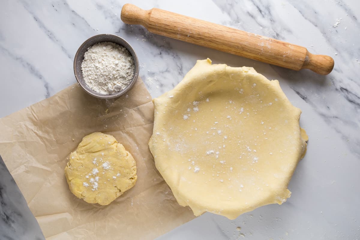 Homemade sourdough pie crust dough over a pie pan with additional dough disc, rolling pin, and bowl of flour nearby.