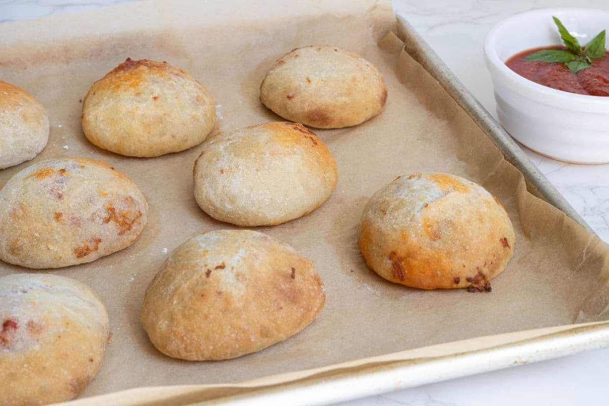 Freshly baked sourdough pizza pockets cooling on a baking tray with a bowl of marinara sauce nearby.