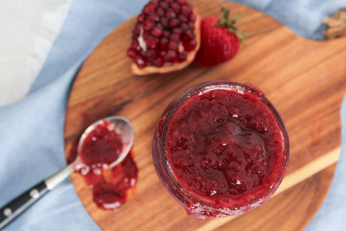 Overhead view of strawberry pomegranate jam showing chunky fruit texture in a glass jar beside a spoon filled with jam.