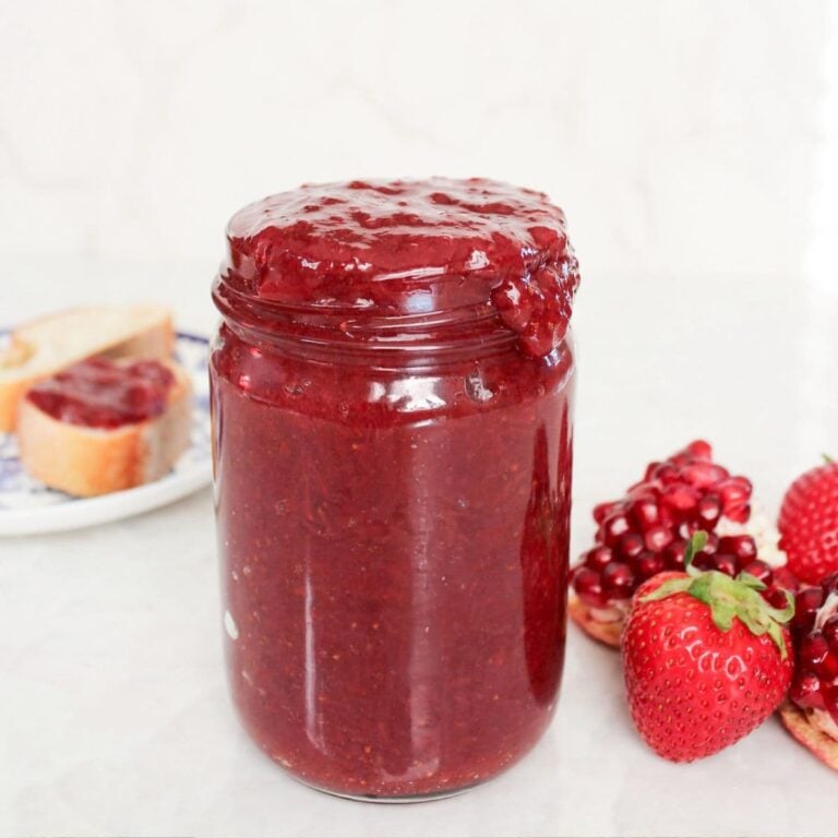 Jar of homemade strawberry pomegranate jam with thick, glossy texture, surrounded by fresh strawberries and pomegranate arils.