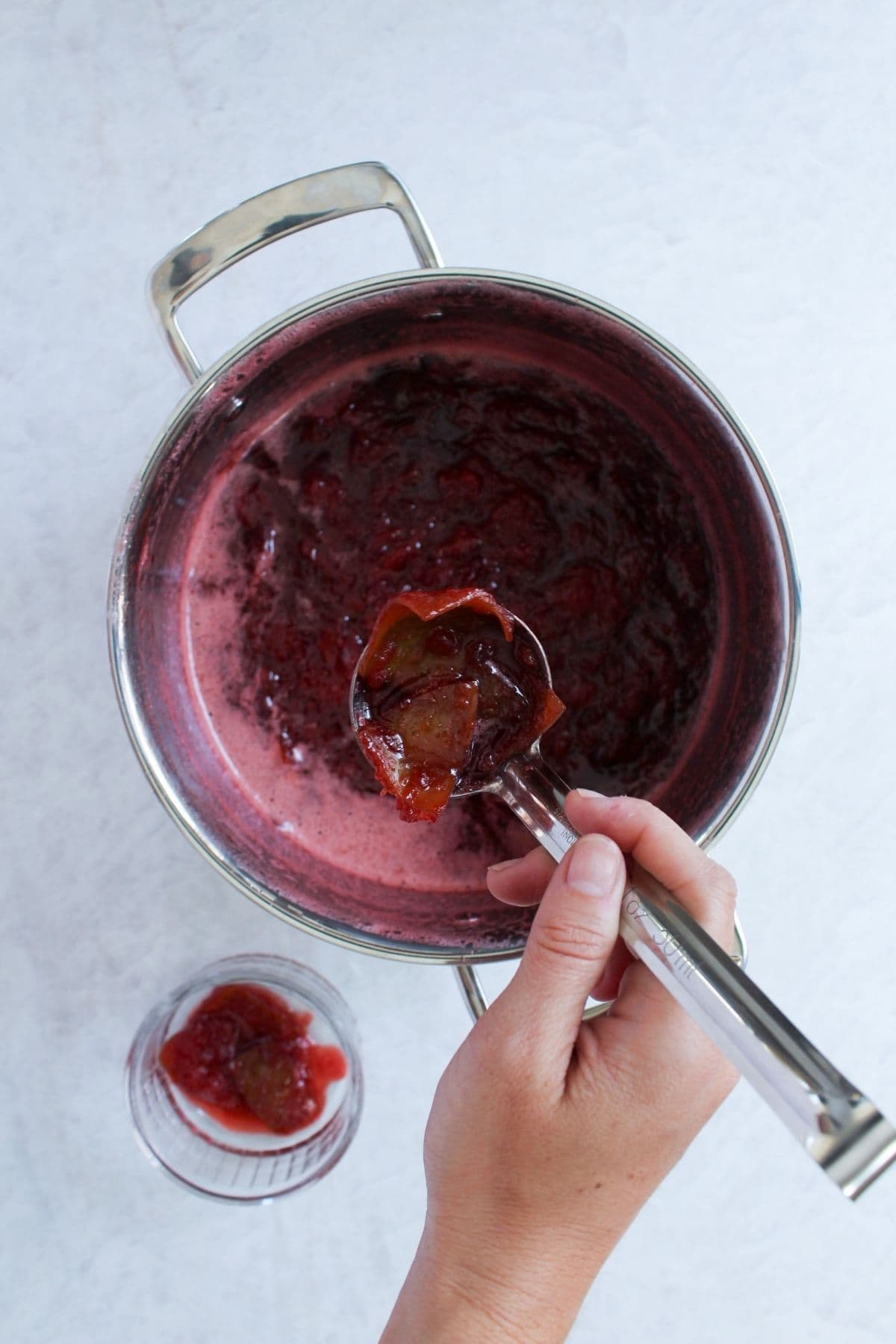 Ladle scooping cooked strawberry pomegranate jam from a saucepan while filling a glass jar for storage.