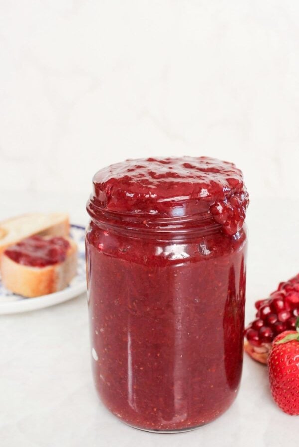 Tall jar of homemade strawberry pomegranate jam with fresh strawberries, pomegranate, and toast in the background.