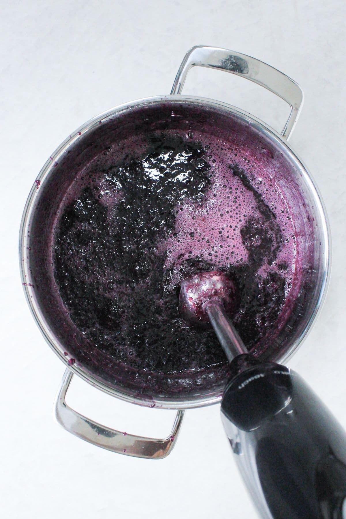 Immersion blender pureeing cooked grapes into a smooth grape jelly mixture in a saucepan.