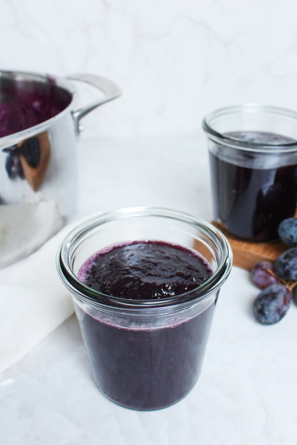 Jar of homemade grape jelly in the foreground with another jar and fresh grapes in the background, next to the pot used for cooking the jelly.