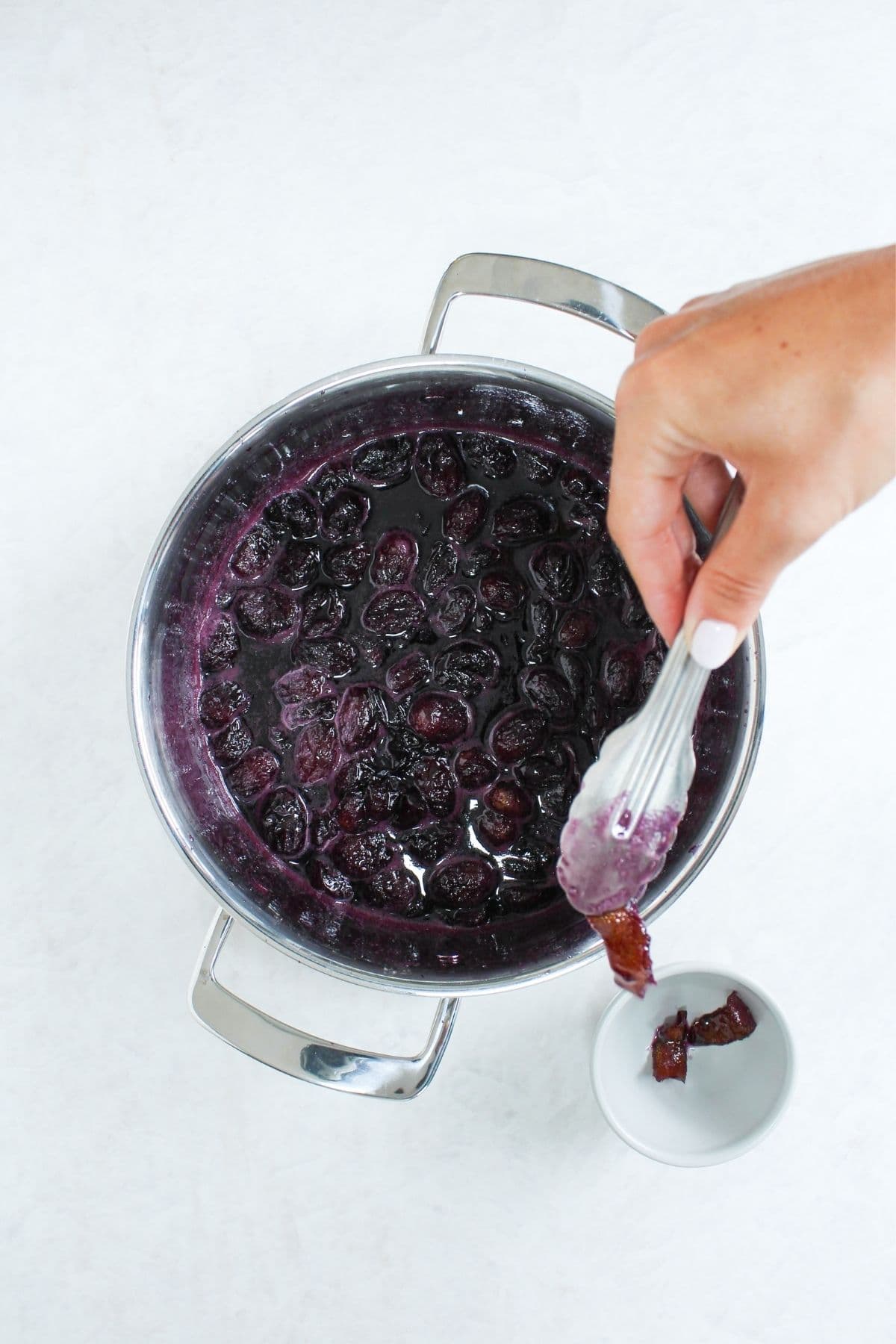 Hand using tongs to remove lemon peel from a pot of cooked grapes before blending.