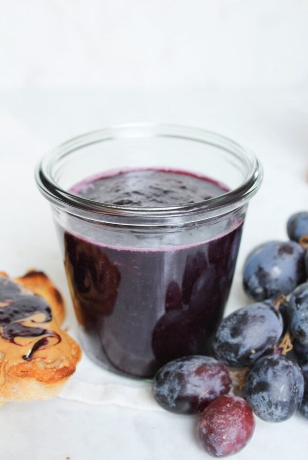 Jar of homemade grape jelly with fresh grapes and peanut butter toast on a countertop.