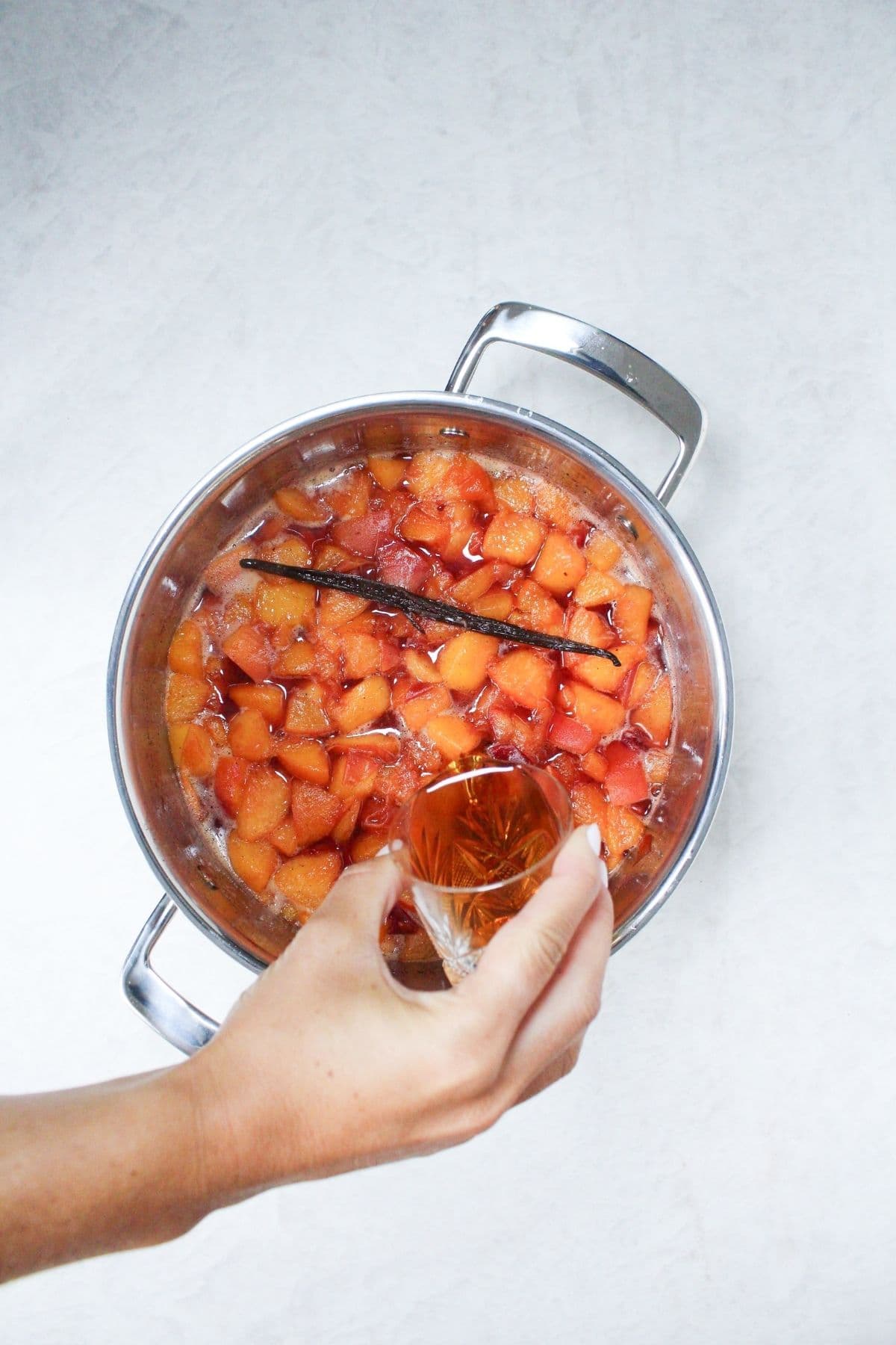 Pouring bourbon into simmering peach vanilla jam with fresh peaches and a whole vanilla bean.