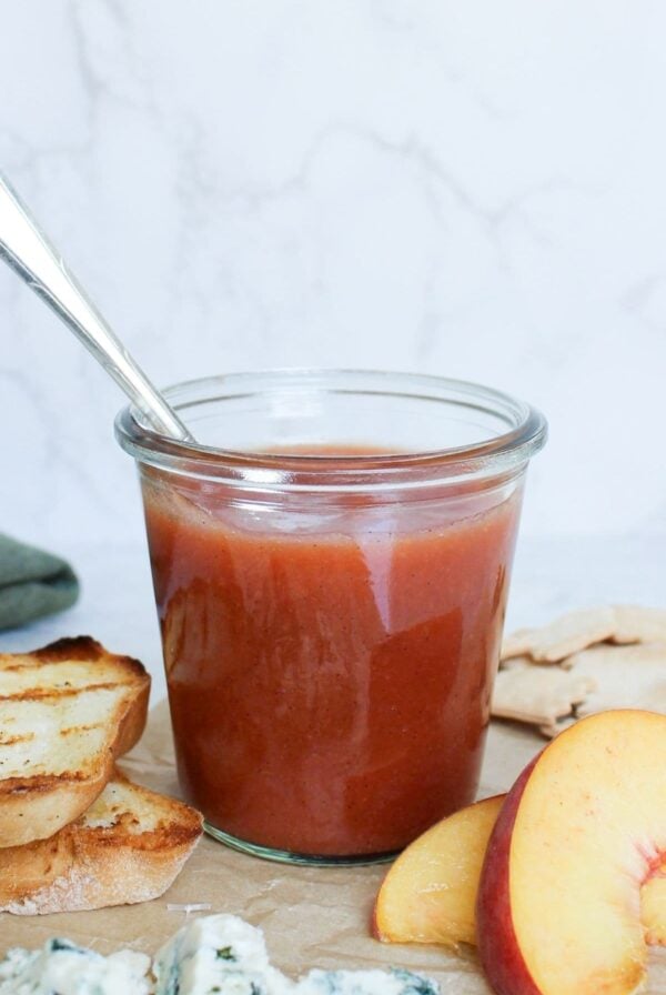 Homemade peach vanilla bourbon jam in a glass jar served with grilled sourdough bread, peaches, sourdough crackers, and blue cheese.
