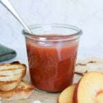 Homemade peach vanilla bourbon jam in a glass jar with grilled bread, fresh peaches, crackers, and blue cheese.
