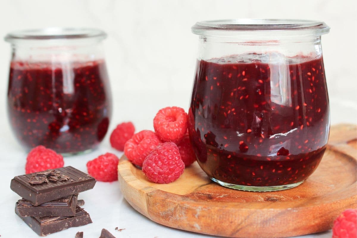 Homemade chocolate raspberry jam in glass jars with fresh raspberries and dark chocolate, made with just three simple ingredients.