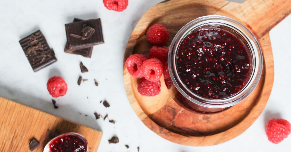 Overhead view of homemade chocolate raspberry jam in a glass jar with fresh raspberries and chopped dark chocolate.