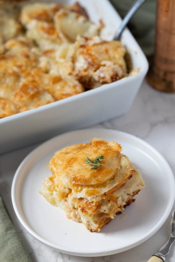 A serving of sourdough au gratin potatoes on a plate with the baking dish behind it.