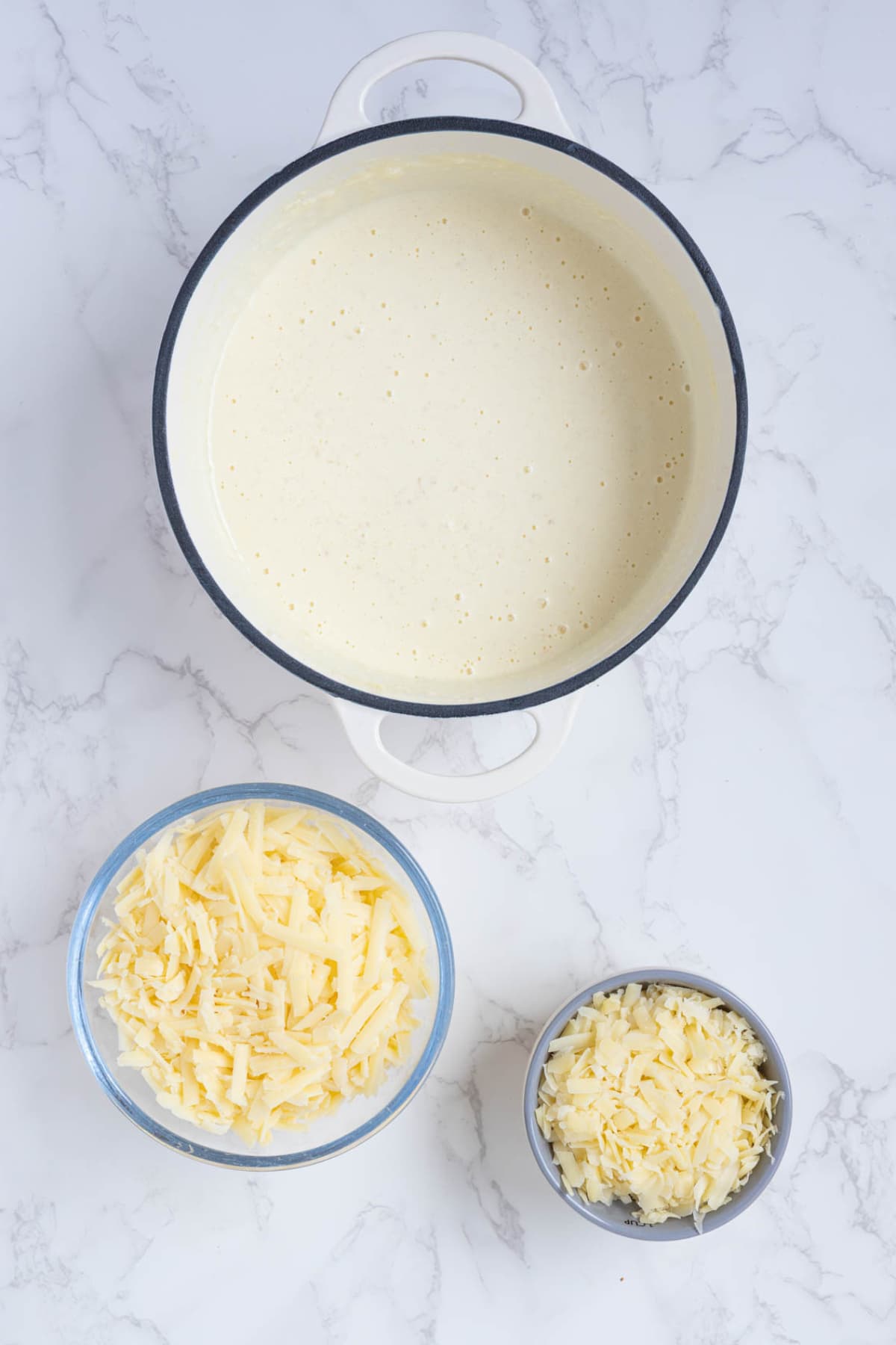 Creamy sourdough cheese sauce in a pot with bowls of shredded cheese nearby.
