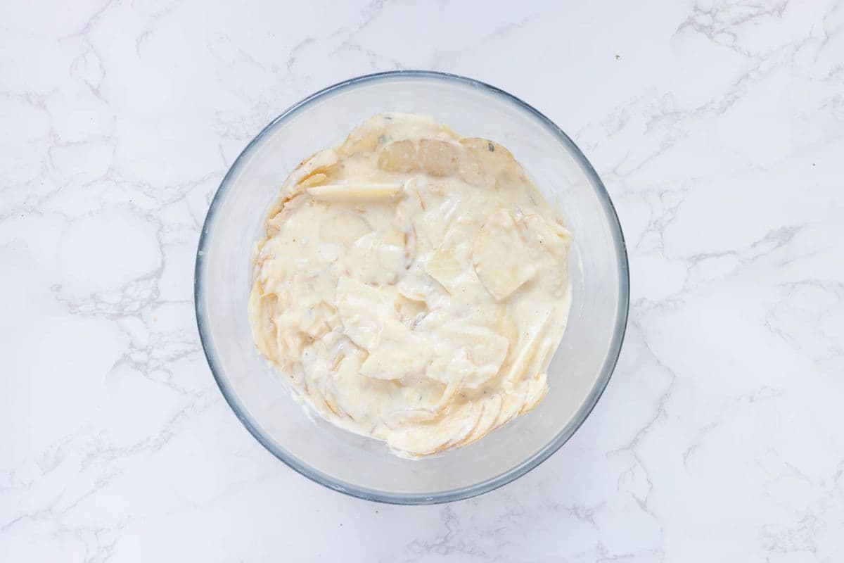 Thinly sliced potatoes coated in sourdough cheese sauce in a mixing bowl before baking.