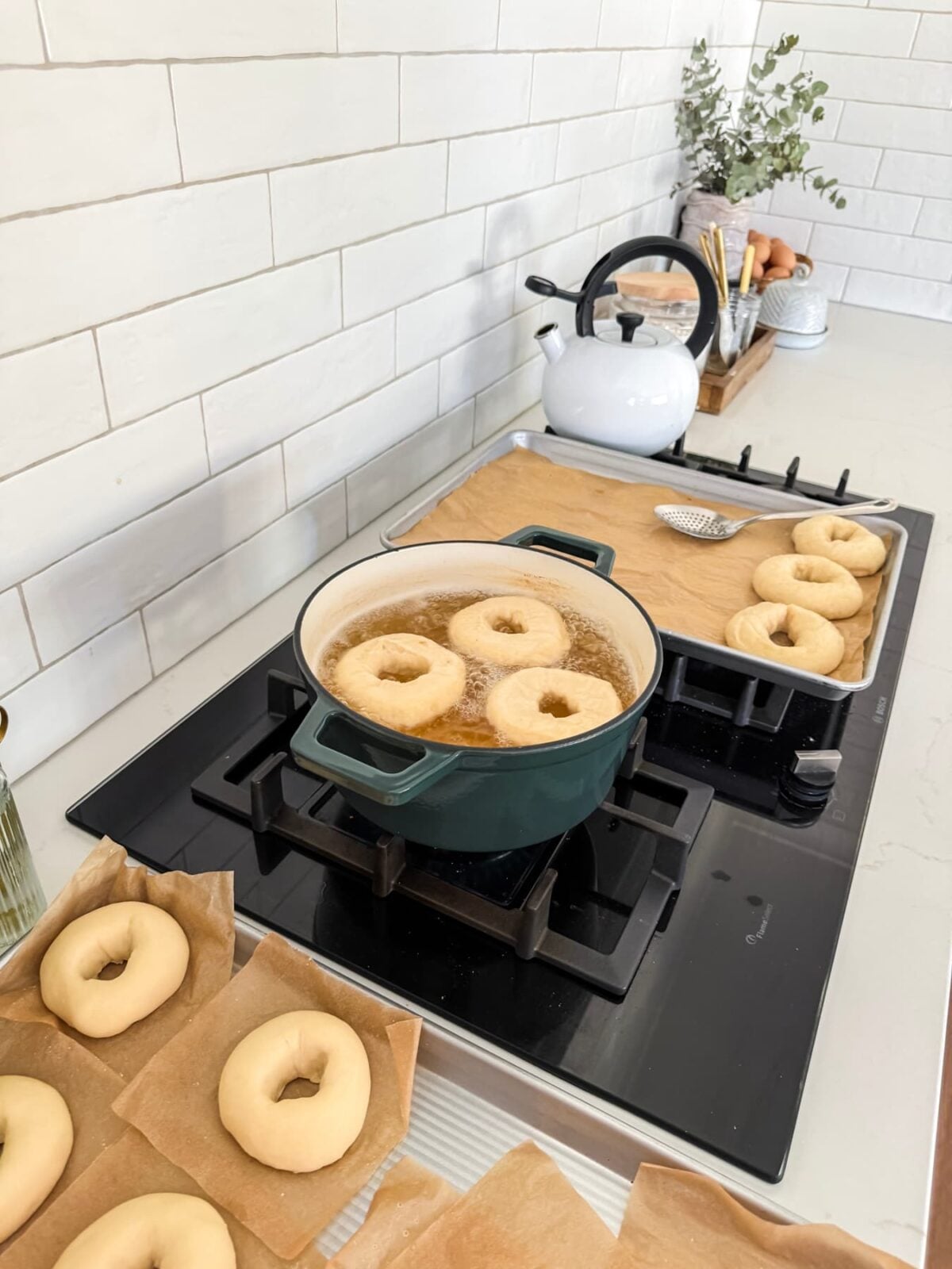 Proofed sourdough bagels on parchment squares beside a pot of boiling honey water with bagels already cooking.