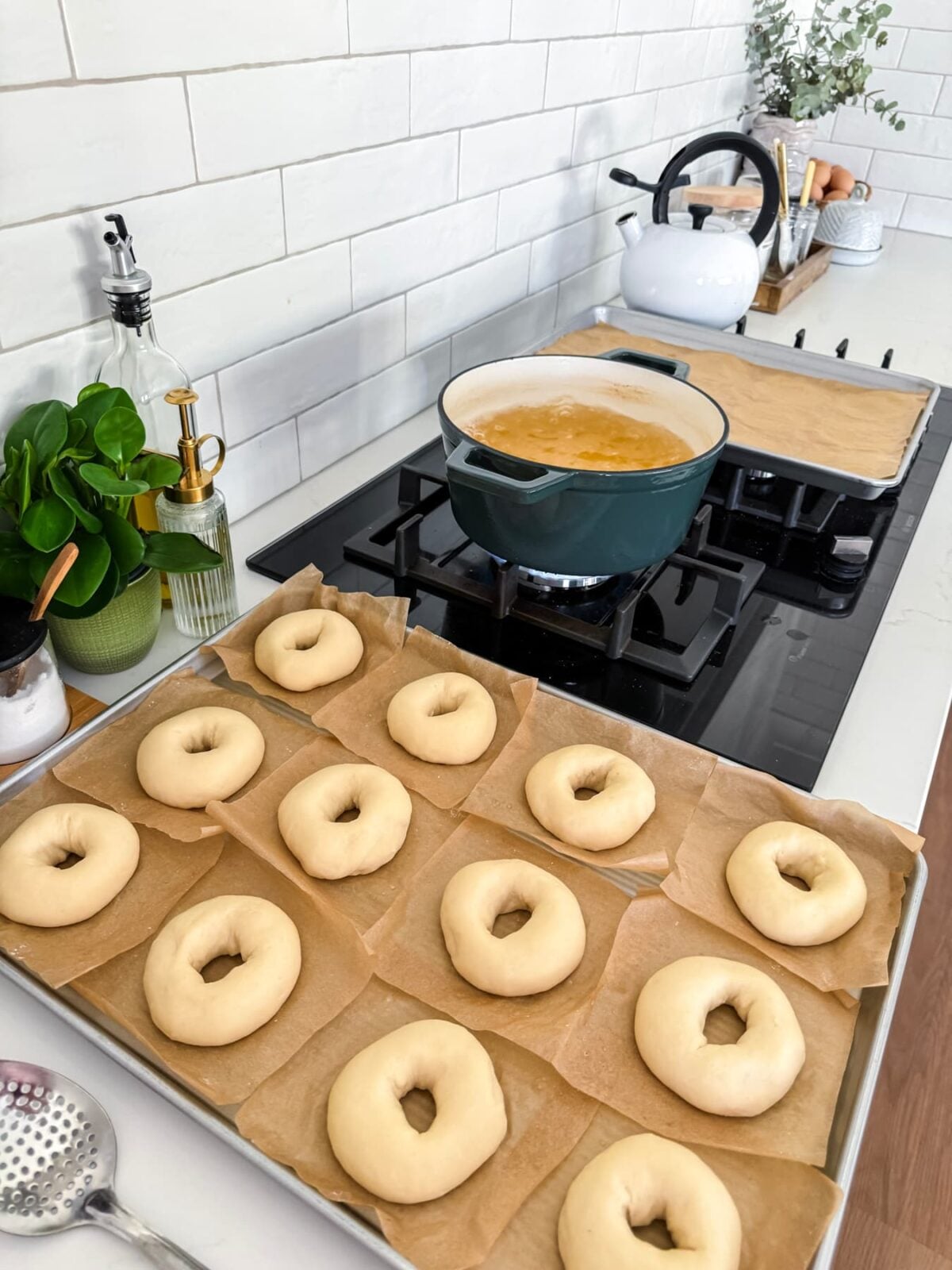 Shaped sourdough bagels proofing on parchment squares beside a pot of honey water ready for boiling.