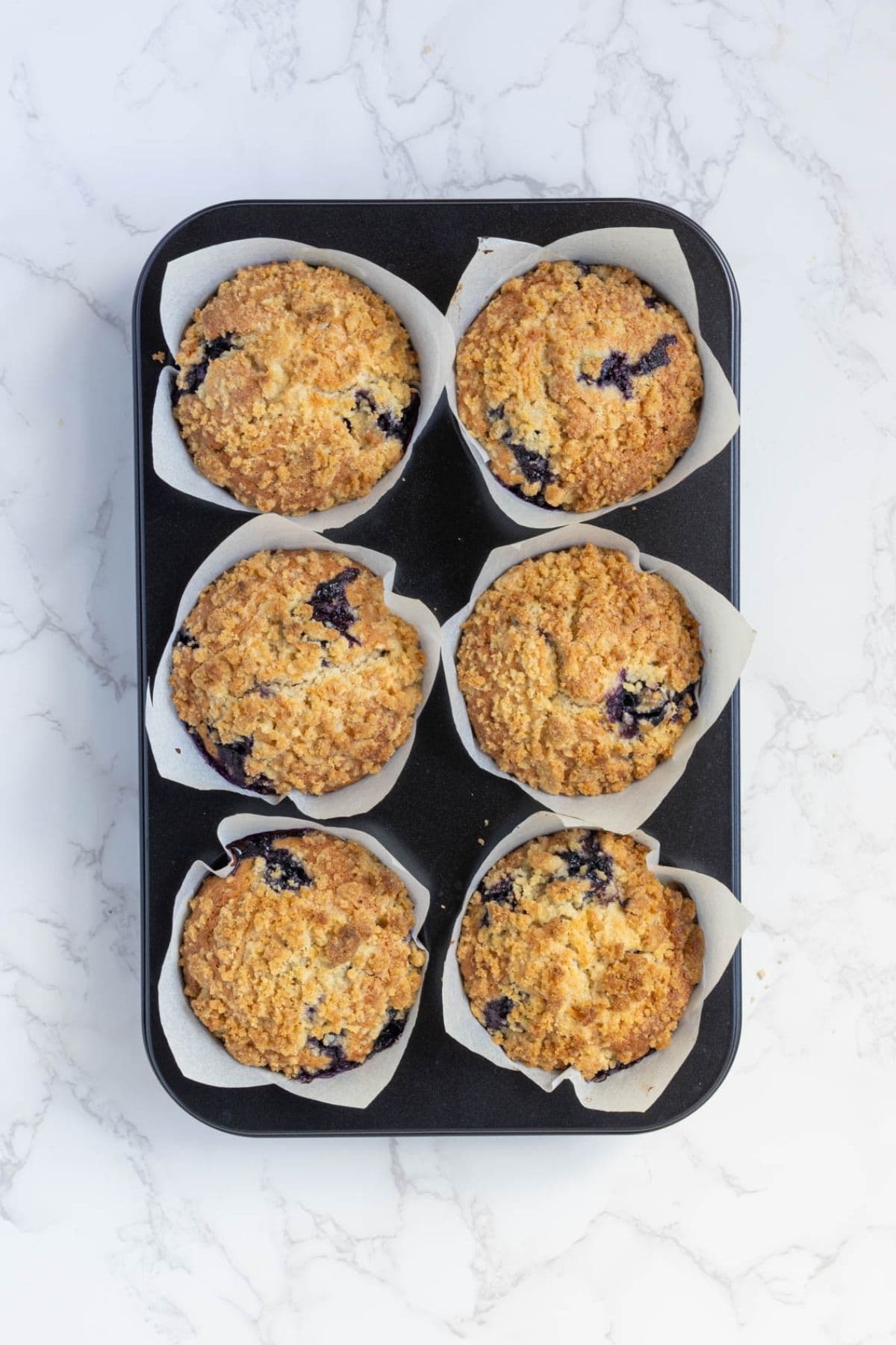 Freshly baked sourdough blueberry muffins with streusel topping cooling in a muffin pan lined with parchment paper.