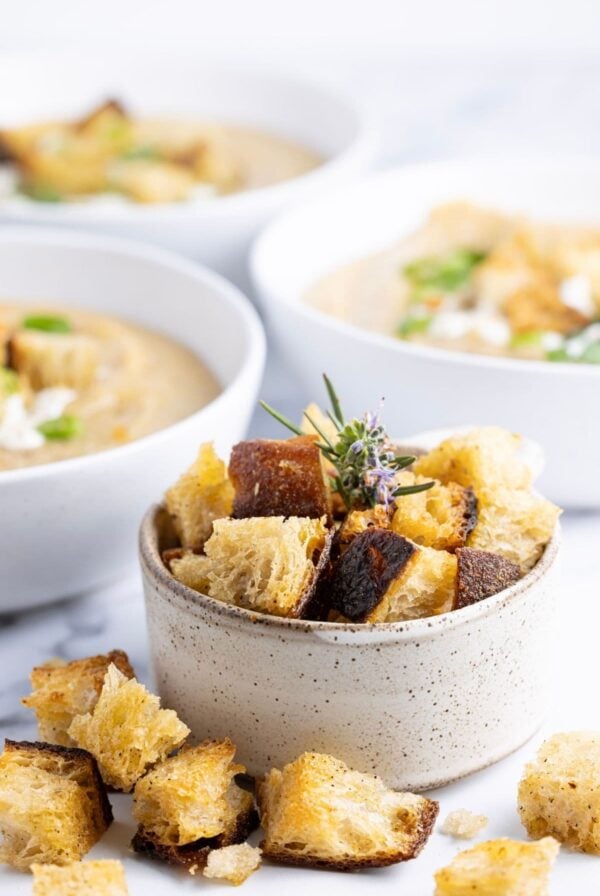 A vertical image of a small speckled dish filled with golden brown sourdough croutons. The croutons are spilling out onto the counter. In the background you can see 3 bowls of vegetable soup, all topped with sourdough croutons and green onions.